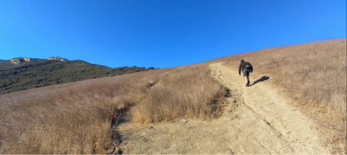 A person walks up a winding dirt path through dry grass and hills under a clear blue sky.