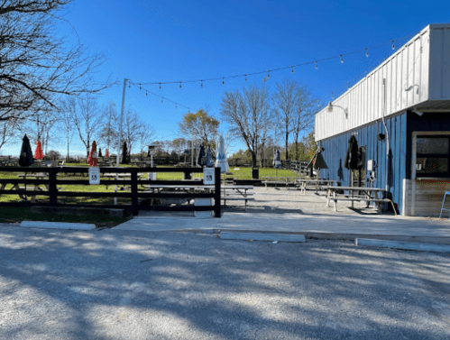 Outdoor seating area with picnic tables, umbrellas, and string lights beside a blue building on a sunny day.