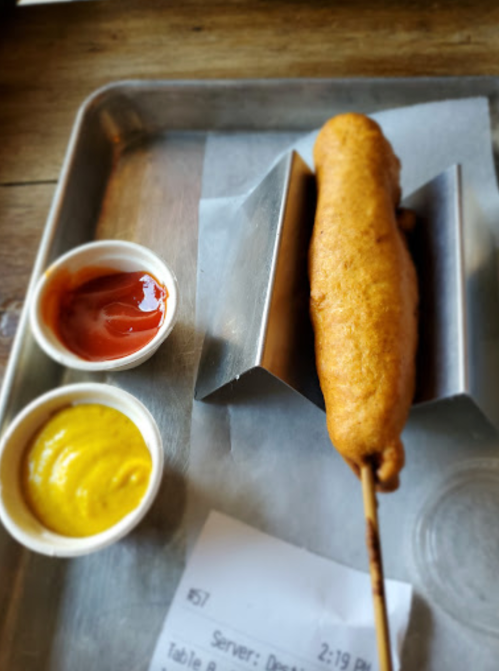A golden-brown corn dog on a tray with ketchup and mustard in small cups beside it.