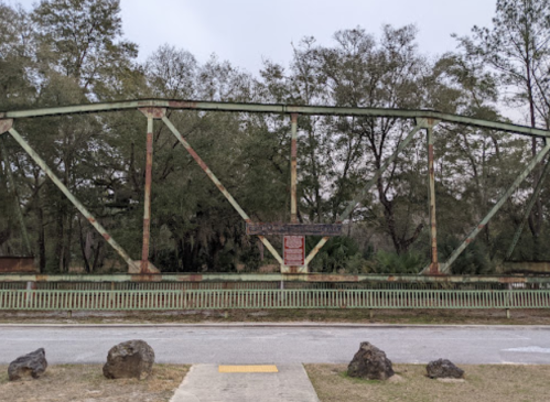 An old, rusted bridge with green metal beams, surrounded by trees and a gravel path in the foreground.