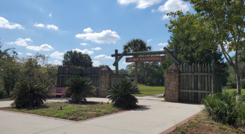 Entrance to Fort Fanning, featuring wooden gates, palm trees, and a clear blue sky with scattered clouds.