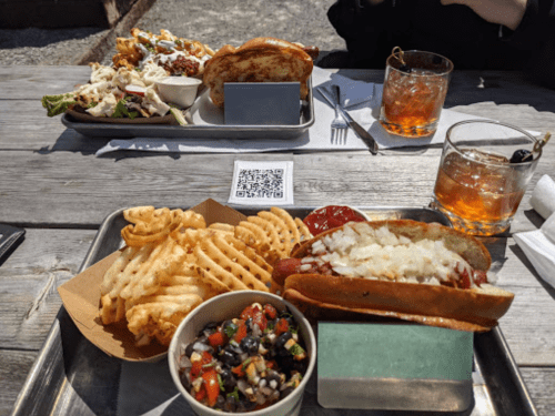 A table with a hot dog, waffle fries, a salad, and two drinks in glasses, set outdoors on a sunny day.