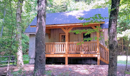 A cozy wooden cabin surrounded by trees, featuring a porch with a railing and a peaceful forest setting.