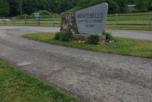 Sign for Montebello Camping and Fishing Resort at a gravel entrance, surrounded by greenery and a wooden fence.