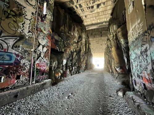 A graffiti-covered tunnel with rocky walls, leading to a bright light at the end, surrounded by gravel.
