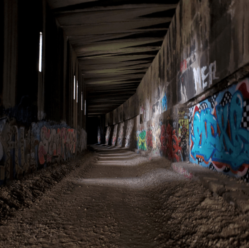 A dimly lit tunnel with curved walls, featuring colorful graffiti and a gravel floor.
