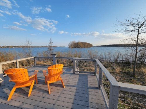 Two bright orange chairs on a wooden deck overlooking a calm lake and distant trees under a blue sky.
