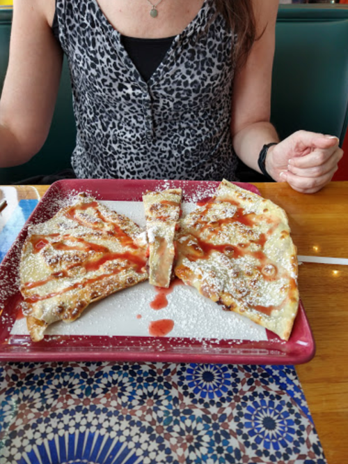 A person in a leopard print top holds a plate of crepes drizzled with sauce and dusted with powdered sugar.