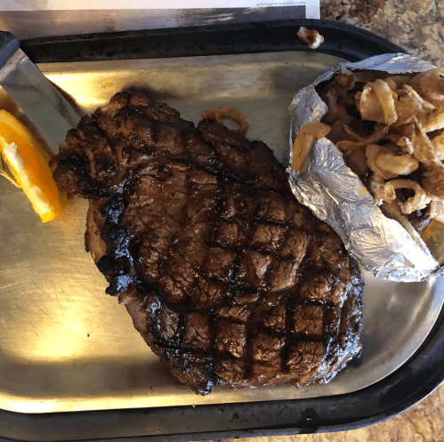 A grilled steak with a charred surface, served on a metal tray alongside a baked potato and crispy onion rings.