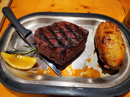 A grilled steak with grill marks, a baked potato, and a lemon wedge on a metal platter.