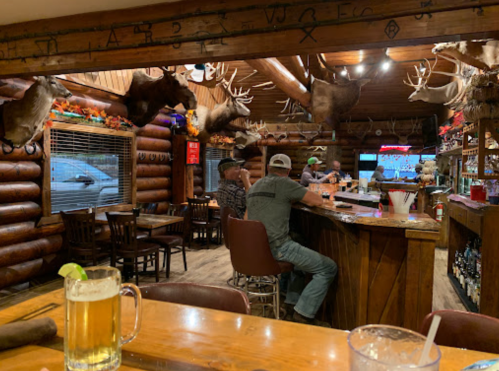 A rustic bar with wooden decor, patrons sitting at the counter, and mounted animal heads on the walls.