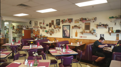 Cozy diner interior with purple booths, tables set with flowers, and vintage decor on the walls. A customer enjoys a meal.
