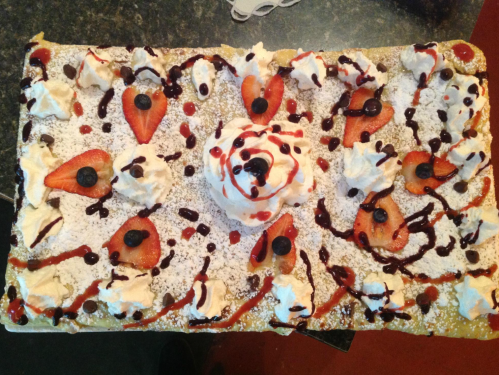 A large dessert topped with whipped cream, strawberries, chocolate drizzle, and decorative patterns on a flat surface.
