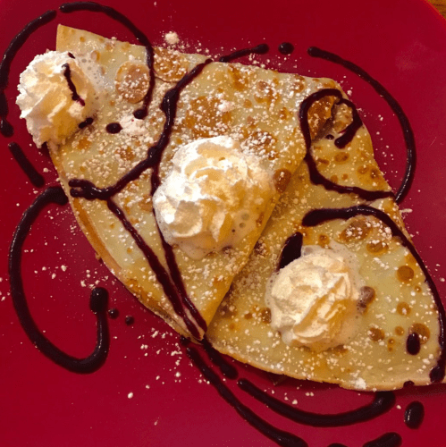 Two crepes topped with whipped cream and drizzled with chocolate sauce, served on a red plate with powdered sugar.
