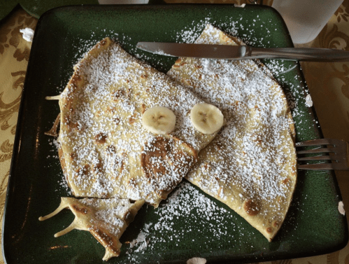 A plate of crepes dusted with powdered sugar and topped with banana slices, served with a knife and fork.