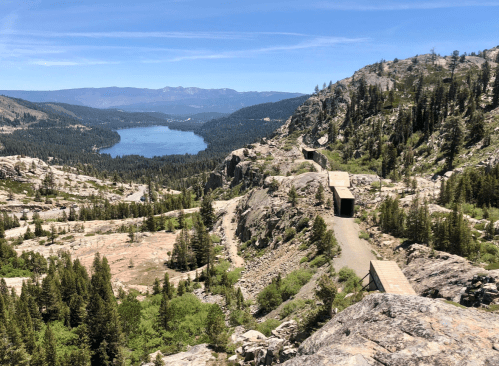 A scenic view of a mountainous landscape with a lake, surrounded by trees and rocky terrain under a clear blue sky.