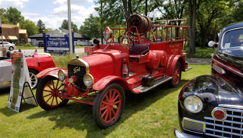 A vintage red fire truck parked on grass, surrounded by trees and other classic cars in a sunny outdoor setting.