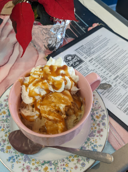A pink cup filled with dessert topped with whipped cream and caramel sauce, next to a menu and a poinsettia plant.