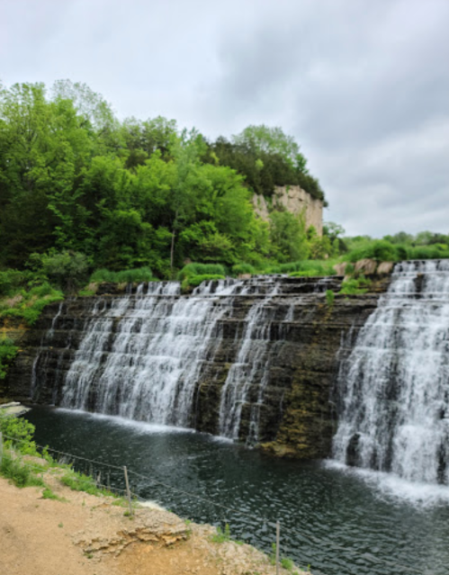 A scenic view of a waterfall cascading over rocky cliffs, surrounded by lush green trees under a cloudy sky.