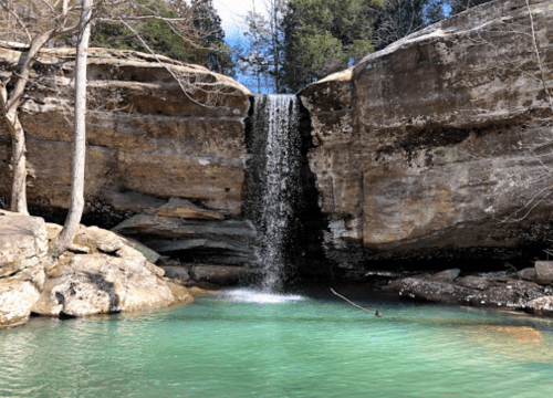 A serene waterfall cascades over rocky cliffs into a clear, turquoise pool surrounded by trees.