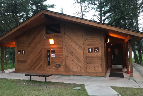 A wooden restroom building with accessible signs, surrounded by trees and grass, under a cloudy sky.