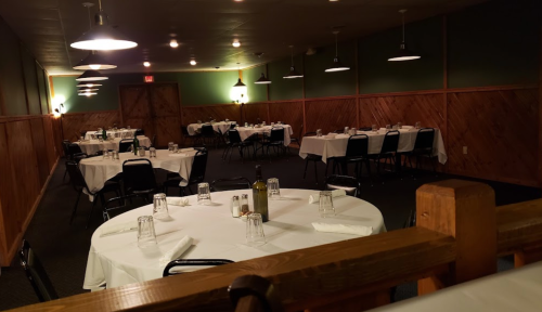 A dimly lit restaurant with empty tables set for dining, featuring white tablecloths and wooden accents.
