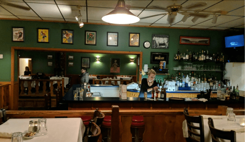 A cozy bar with a green wall, featuring a bartender, shelves of drinks, and framed artwork. Tables are set for dining.