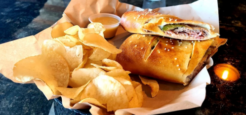 A plate with a golden-brown sandwich filled with meat and pickles, accompanied by a side of potato chips and dipping sauce.