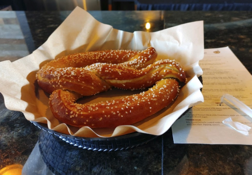 A basket with two twisted pretzels topped with sesame seeds, resting on a paper liner on a dark countertop.