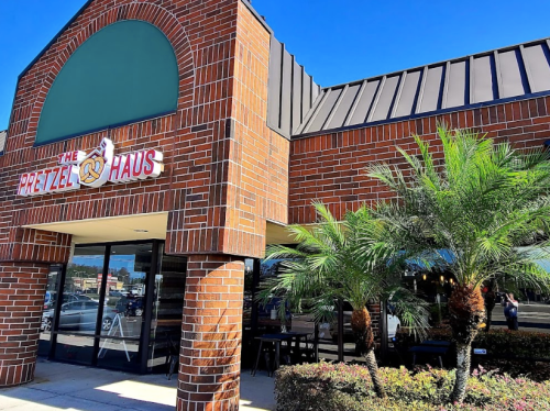 Exterior of The Pretzel Haus, featuring a brick facade, palm trees, and a clear blue sky.