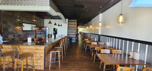 Interior of a modern restaurant featuring wooden tables, bar seating, and warm lighting along a long, narrow space.