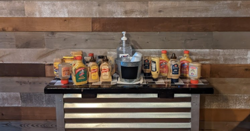 A table displaying various condiment bottles, including mustards and sauces, against a wooden wall backdrop.