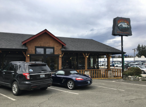 Exterior of a restaurant with a wooden facade, a sign, and parked cars in the foreground under a cloudy sky.