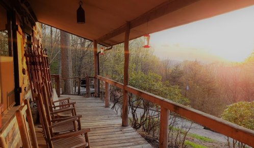 A wooden porch with rocking chairs overlooks a sunset through trees, creating a serene, rustic atmosphere.