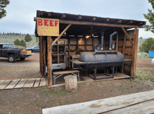A rustic outdoor food stall with a large smoker, wooden structure, and a sign that reads "BEEF." Truck parked nearby.