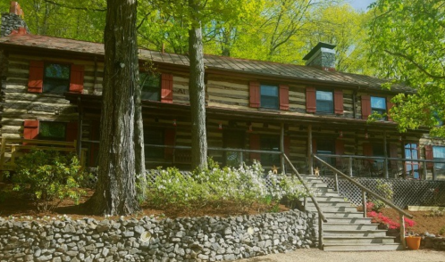 A rustic log cabin with red shutters, surrounded by greenery and a stone pathway leading to the entrance.