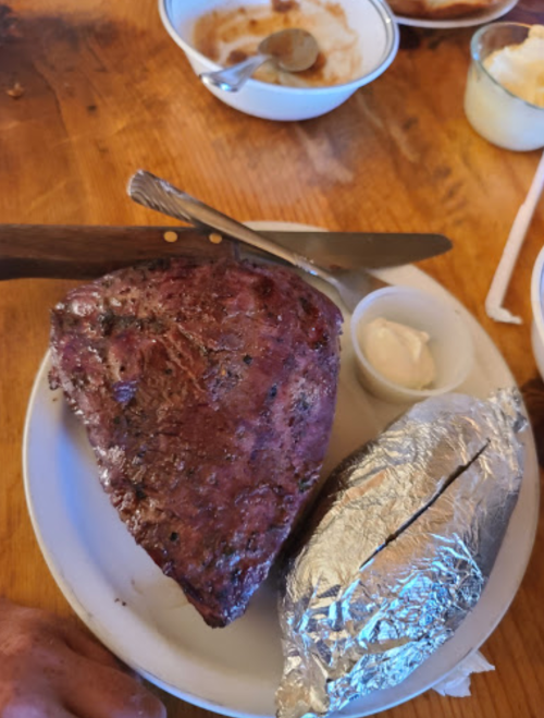 A plate with a large piece of grilled meat and a foil-wrapped baked potato, accompanied by a small dish of sour cream.