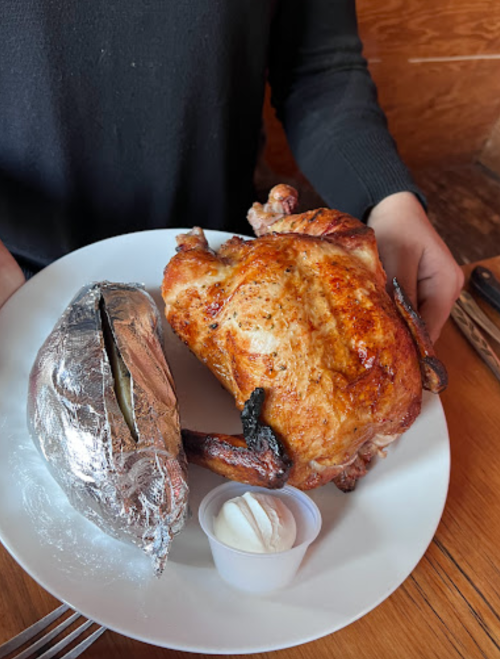 A person holds a plate with a whole roasted chicken and a baked potato wrapped in foil, served with sour cream.