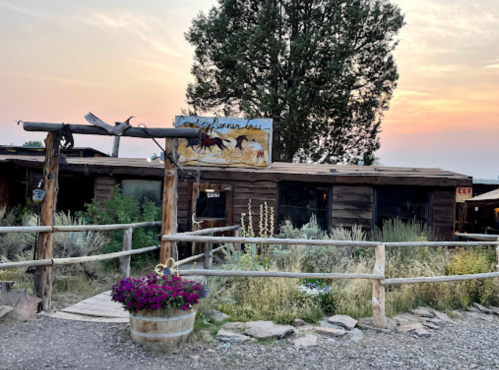 A rustic wooden building with a horse-themed sign, surrounded by greenery and flowers, under a sunset sky.