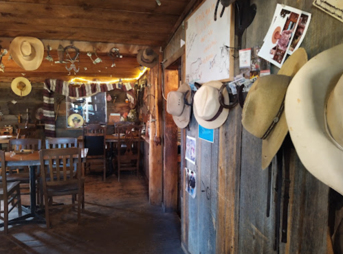 Interior of a rustic restaurant with wooden walls, cowboy hats hanging, and a cozy dining area visible.