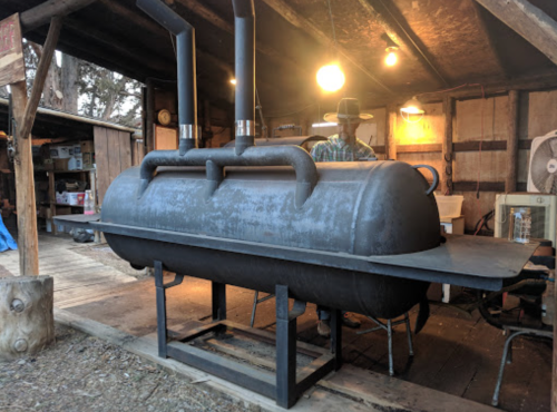 A large, black smoker sits in a rustic wooden shed, with a person working in the background under warm lighting.