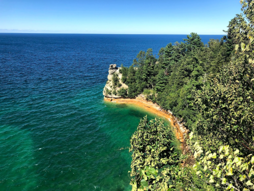 A scenic view of a rocky coastline meeting a vibrant blue lake, surrounded by lush green trees under a clear sky.
