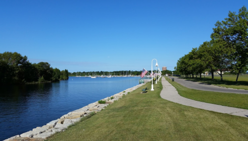 A serene waterfront scene with a clear blue sky, boats in the distance, and a pathway lined with trees.