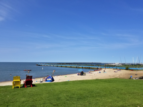 A sunny beach scene with a pier, boats, and colorful chairs on the grass, inviting relaxation and outdoor activities.