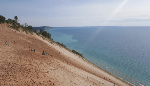 A sandy dune slopes down to a calm blue lake, with people enjoying the scenery and climbing the hill.