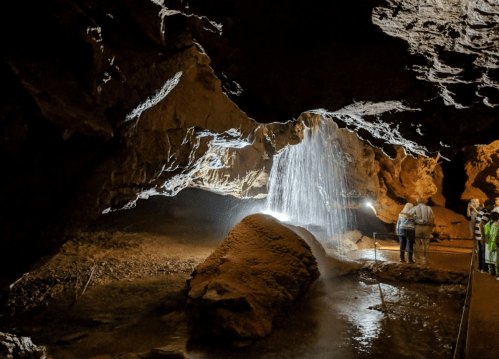 A cave interior with a waterfall, illuminated by lights, and a person standing near a large rock formation.