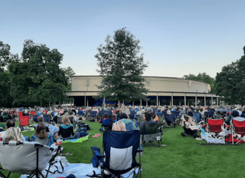 A large crowd sits on blankets and chairs in a park, facing a stage surrounded by trees during an outdoor event.