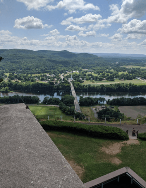 A scenic view from a high vantage point, overlooking a river, bridge, and lush green hills under a partly cloudy sky.