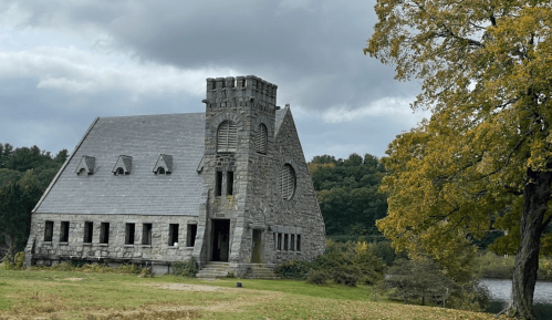An old stone building with a tower, surrounded by trees and a river under a cloudy sky.