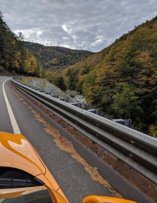 A winding road alongside a river, surrounded by autumn-colored trees and mountains under a cloudy sky.